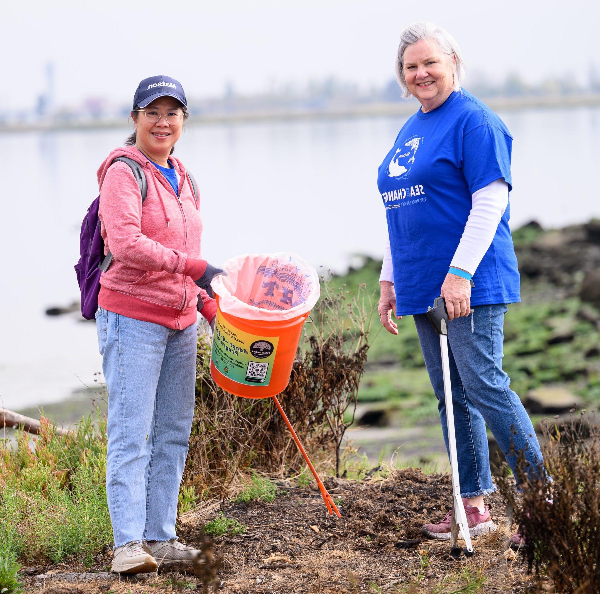 A volunteer holds a grabber while another holds a bucket and grabber to collect trash.